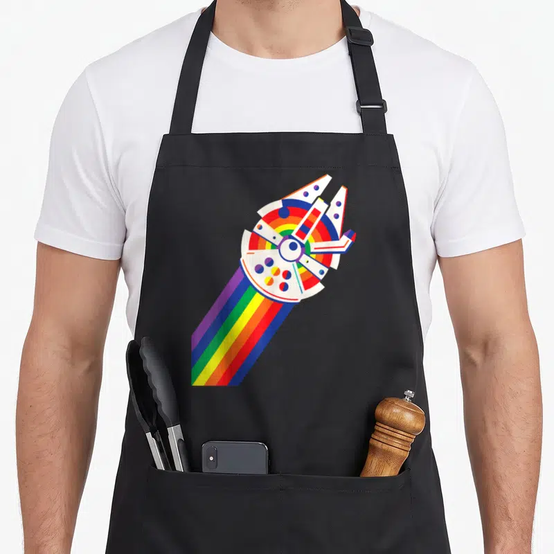 Rainbow falcon apron - Stylized white spaceship with diagonal rainbow trail in red yellow green blue purple — shown on a black cotton canvas apron against a white background