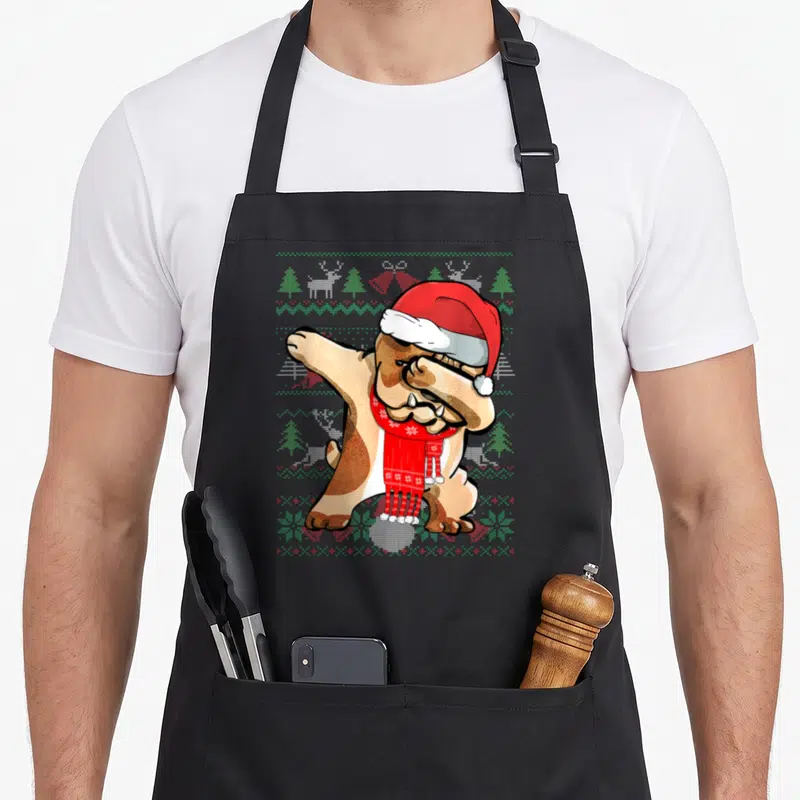 Dabbing ugly sweater apron - Cartoon bulldog dabbing in Santa hat and red scarf on knit pattern background — shown on a black cotton canvas apron against a white background