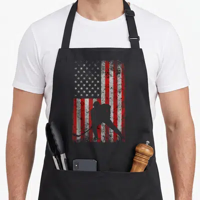 Ice american flag apron - Grey hockey player silhouette over distressed red white and grey US flag pattern — shown on a black cotton canvas apron against a white background