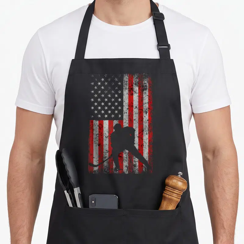 Ice american flag apron - Grey hockey player silhouette over distressed red white and grey US flag pattern — shown on a black cotton canvas apron against a white background