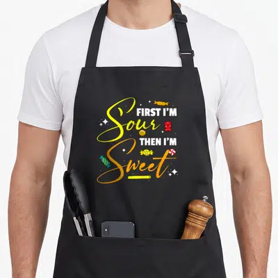 Sour candy aprons - Yellow and orange script reading First I'm Sour Then I'm Sweet with gummy bears and wrapped candies — shown on a black cotton canvas apron against a white background