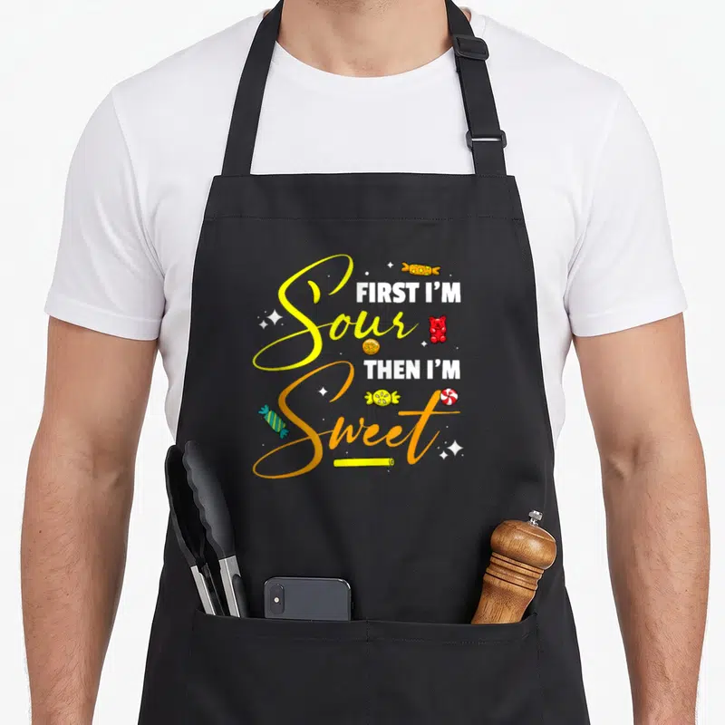 Sour candy aprons - Yellow and orange script reading First I'm Sour Then I'm Sweet with gummy bears and wrapped candies — shown on a black cotton canvas apron against a white background