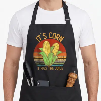 Corn family thanksgiving apron - Retro two corn cobs with arched text IT'S CORN IT HAS THE JUICE on striped sun — shown on a black cotton canvas apron against a white background