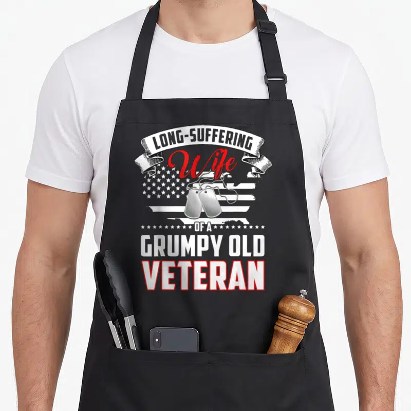 Suffering wife apron - Stacked text LONG-SUFFERING Wife OF A GRUMPY OLD VETERAN with dog tags and distressed flag — shown on a black cotton canvas apron against a white background