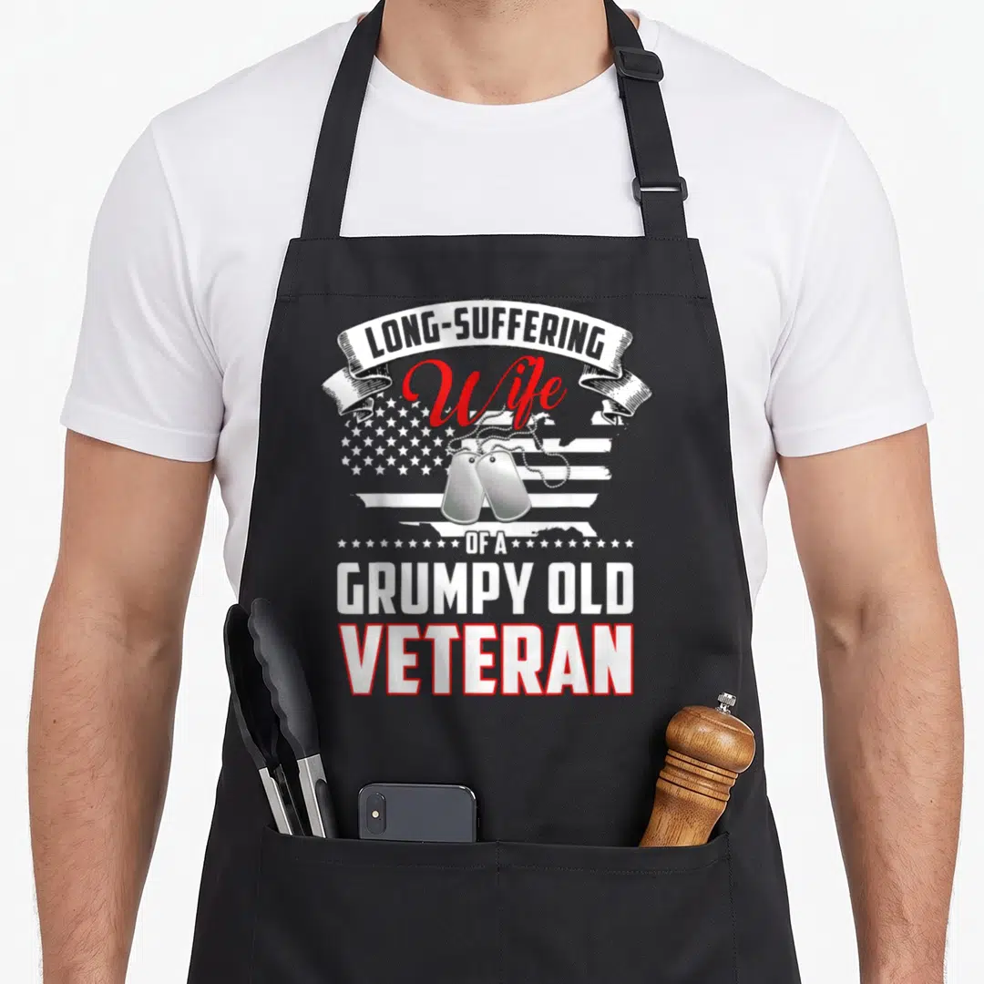 Suffering wife apron - Stacked text LONG-SUFFERING Wife OF A GRUMPY OLD VETERAN with dog tags and distressed flag — shown on a black cotton canvas apron against a white background
