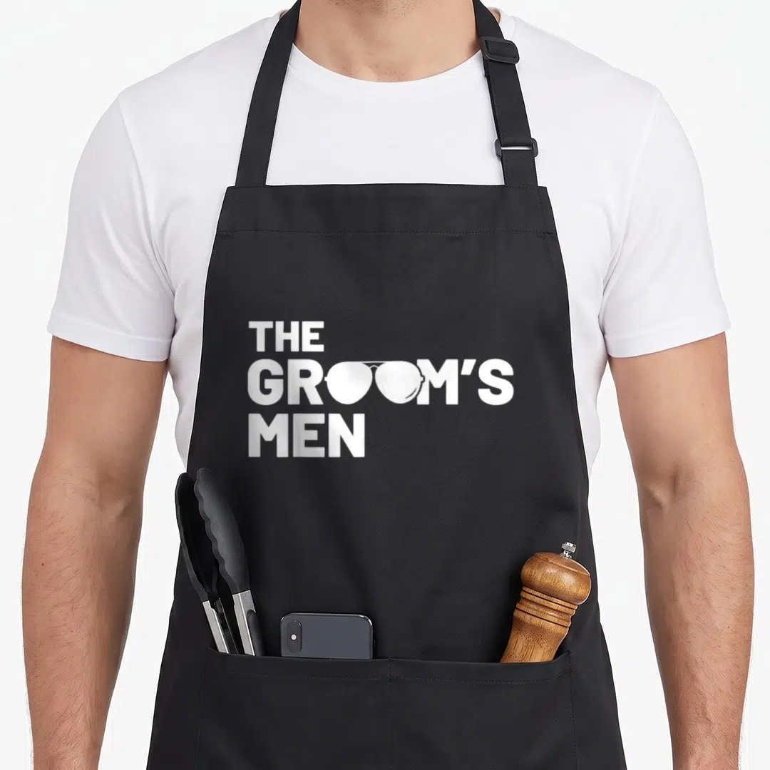 Squat funny wedding aprons - White text THE GROOM'S MEN with sunglasses replacing two O letters in stacked layout — shown on a black cotton canvas apron against a white background