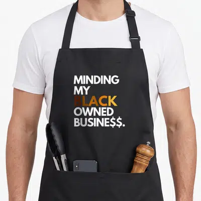 Owned business apron - Stacked text Minding My Black Owned Business in white brown orange yellow with dollar signs — shown on a black cotton canvas apron against a white background