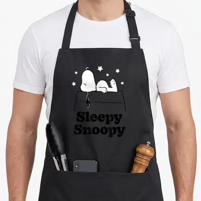 Sleepy peanut lover apron - Snoopy lying on doghouse with stars and stacked Sleepy Snoopy text — shown on a black cotton canvas apron against a white background