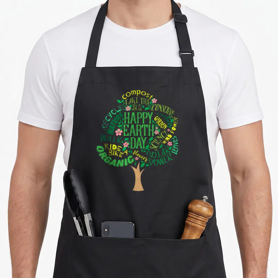Earth tree apron - Tree canopy formed by words compost, solar power, and happy earth day with pink flowers — shown on a black cotton canvas apron against a white background