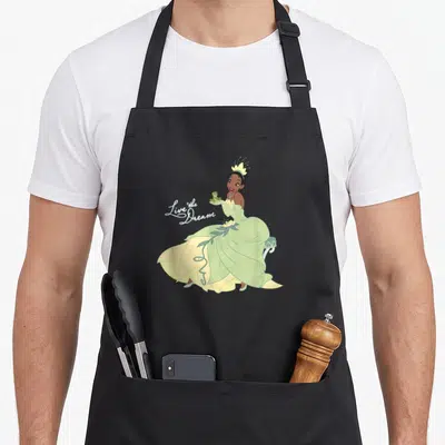 Dream princess apron - Princess in green dress sitting on bench holding frog next to script text — shown on a black cotton canvas apron against a white background