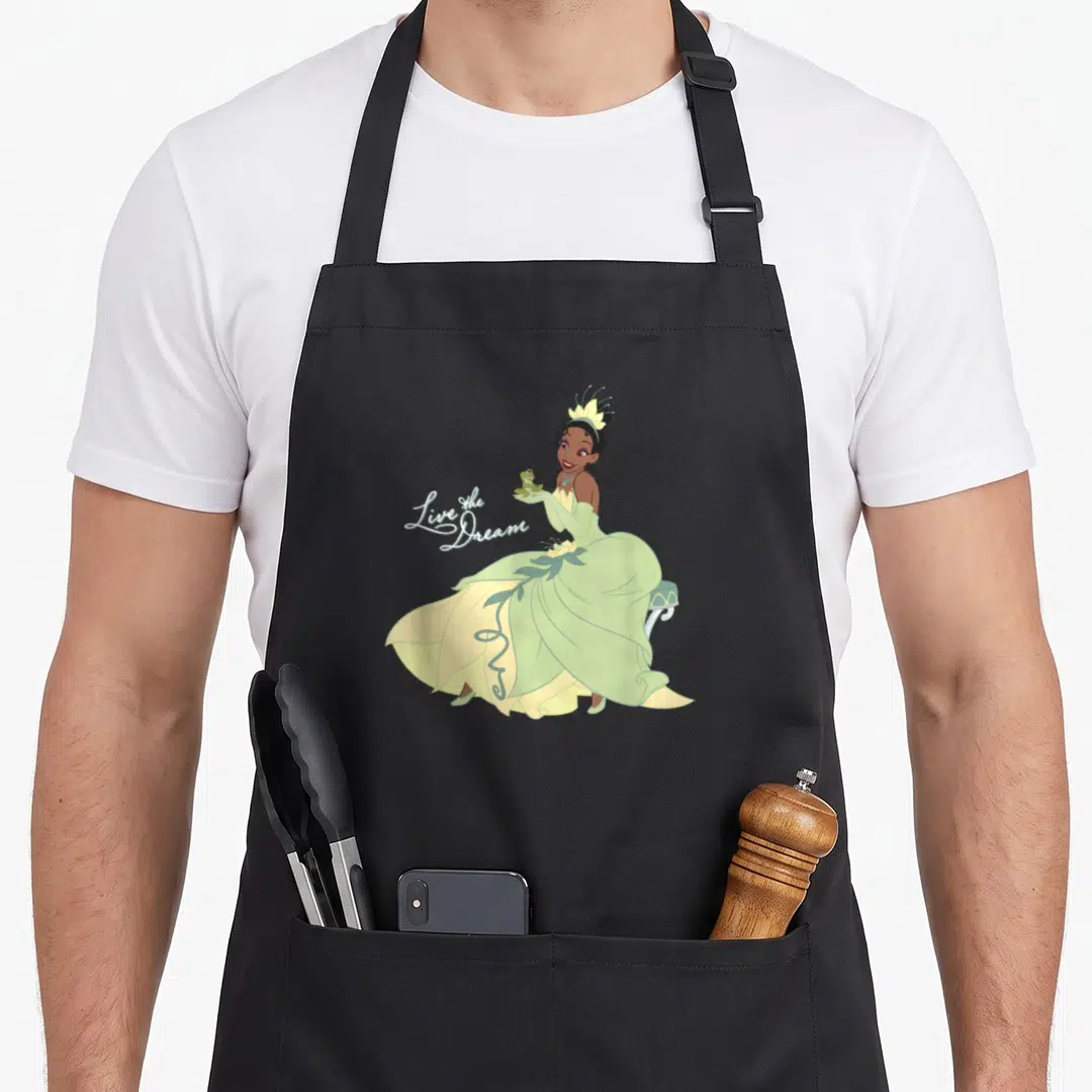 Dream princess apron - Princess in green dress sitting on bench holding frog next to script text — shown on a black cotton canvas apron against a white background