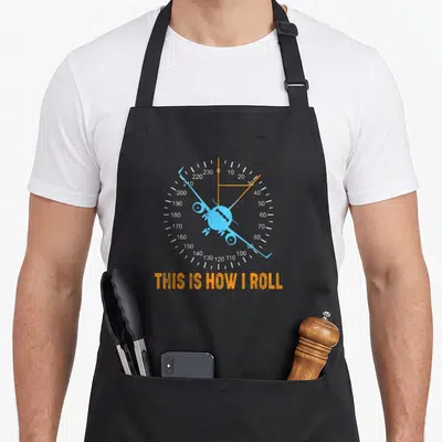 Roll airplane apron - Light blue plane on white dial with orange needle and THIS IS HOW I ROLL text — shown on a black cotton canvas apron against a white background