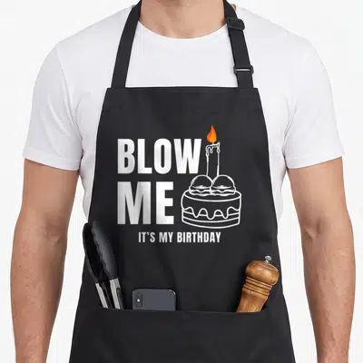 Blow the dirty apron - White stacked blow me text next to birthday cake with orange flame candle — shown on a black cotton canvas apron against a white background