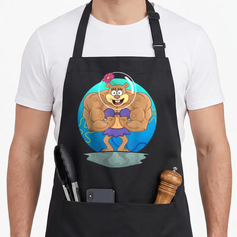 Sandy muscle apron - Cartoon brown bear in purple bikini inside blue circle with pink flower — shown on a black cotton canvas apron against a white background
