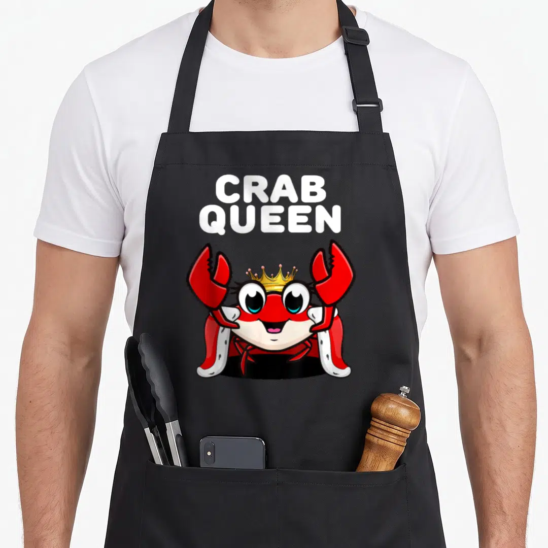 Queen crab apron - Cartoon red crab wearing a yellow crown and red cape under white CRAB QUEEN text — shown on a black cotton canvas apron against a white background