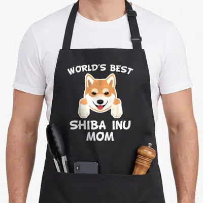 Shiba inu owner dog apron - Cartoon Shiba Inu head with paws, arched text 'World's Best', stacked 'Shiba Inu Mom' — shown on a black cotton canvas apron against a white background
