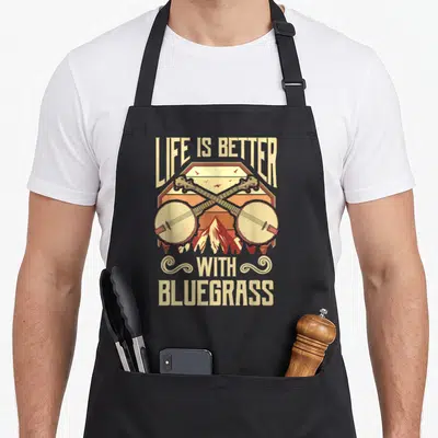 Banjo bluegrass apron - Crossed banjos over mountains with LIFE IS BETTER WITH BLUEGRASS text in retro colors — shown on a black cotton canvas apron against a white background