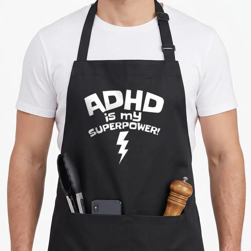 Inspirational adhd apron - White stacked text ADHD is my SUPERPOWER with lightning bolt below — shown on a black cotton canvas apron against a white background