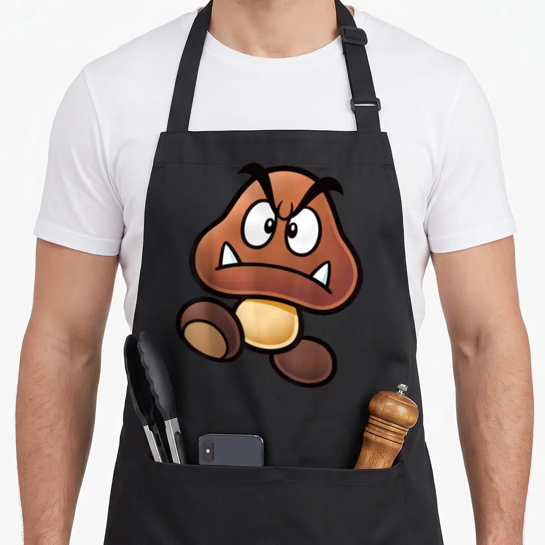 Goomba face apron - Centered brown cartoon mushroom character with angry eyes and fangs — shown on a black cotton canvas apron against a white background