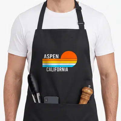 California aspen apron - Retro sun over orange yellow blue stripes with ASPEN CALIFORNIA text — shown on a black cotton canvas apron against a white background
