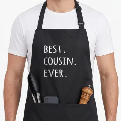 Cousin worlds greatest dad apron - White stacked text reading BEST. COUSIN. EVER on dark background — shown on a black cotton canvas apron against a white background