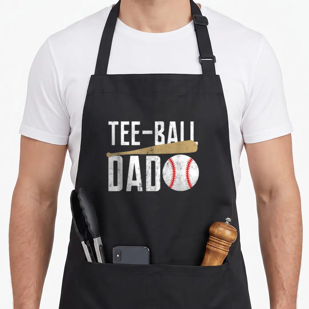 Tball daddy apron - Distressed TEE-BALL DAD text with tan bat and red stitched white baseball graphic — shown on a black cotton canvas apron against a white background