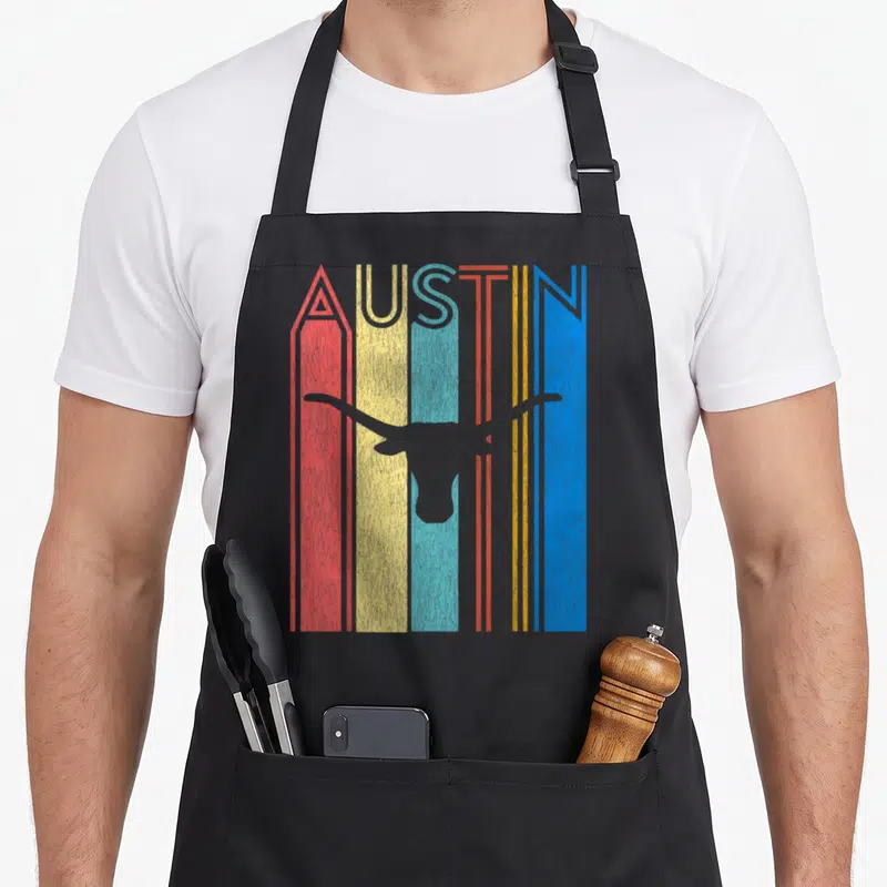 Longhorn texas aprons - Grey longhorn skull silhouette over red yellow teal blue vertical stripes with AUSTIN text — shown on a black cotton canvas apron against a white background