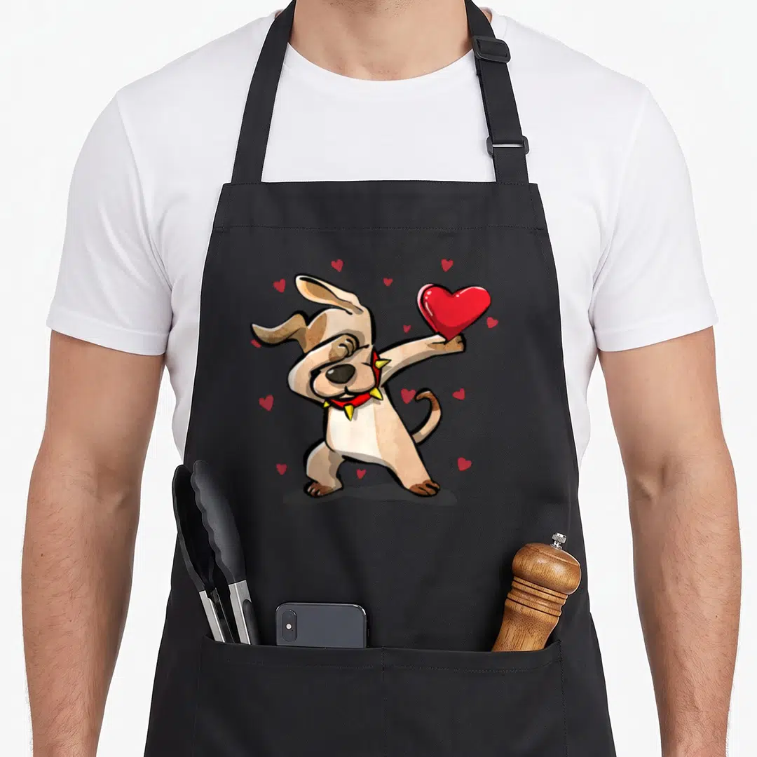 Dabbing dog apron - Cartoon tan dog dabbing while holding a large red heart with smaller hearts scattered around — shown on a black cotton canvas apron against a white background
