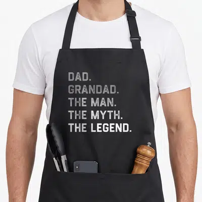 Father grandad aprons - DAD GRANDAD THE MAN THE MYTH THE LEGEND stacked white grey typography — shown on a black cotton canvas apron against a white background