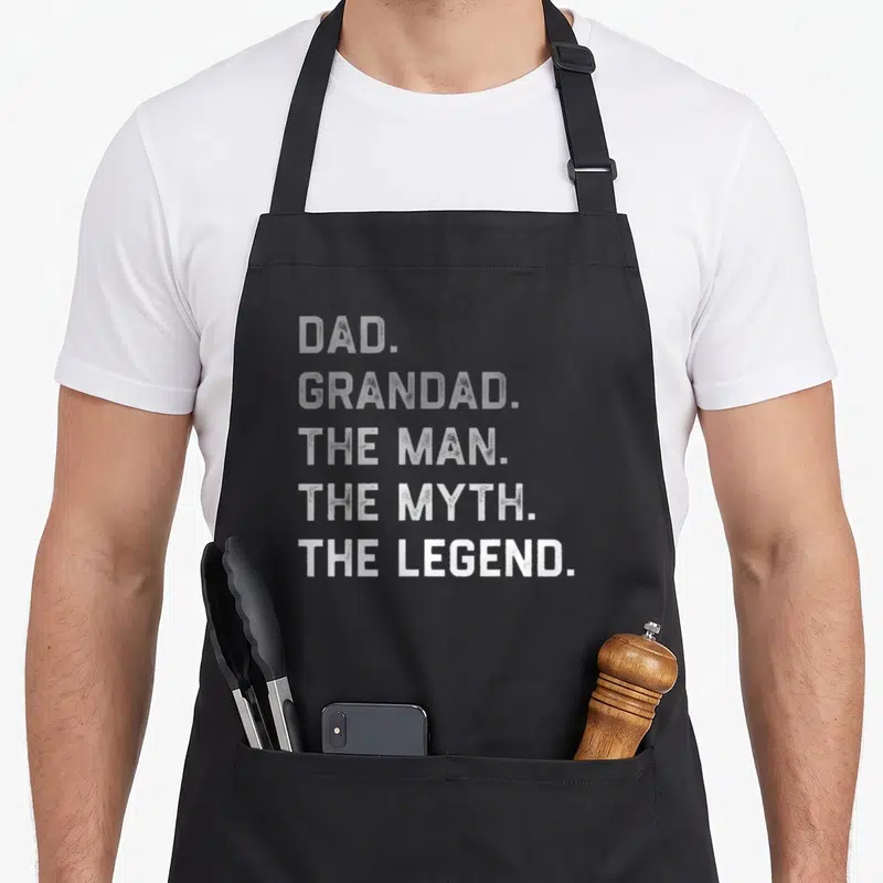 Father grandad aprons - DAD GRANDAD THE MAN THE MYTH THE LEGEND stacked white grey typography — shown on a black cotton canvas apron against a white background