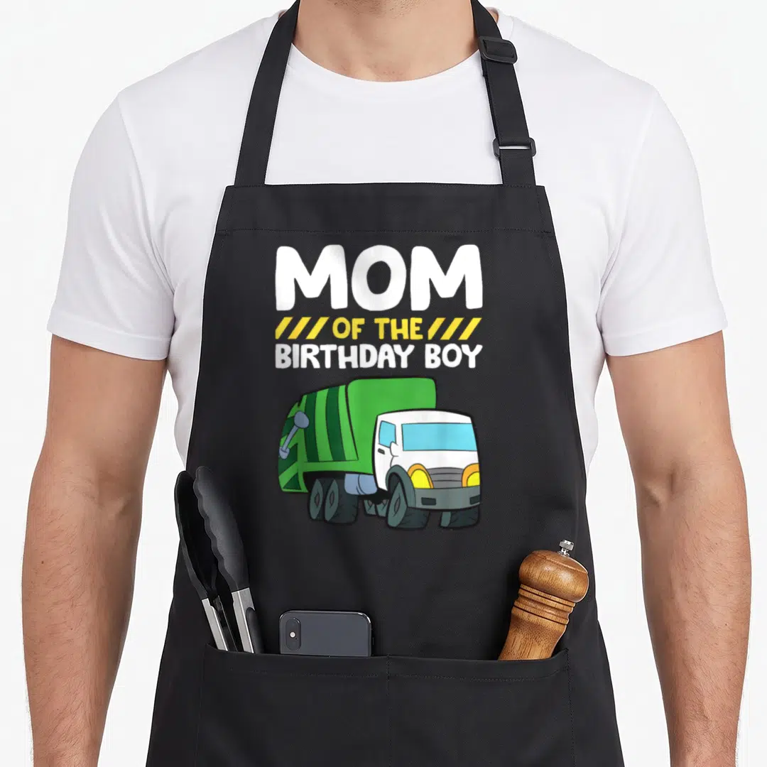Cool garbage truck apron - MOM OF THE BIRTHDAY BOY text above green and white cartoon garbage truck — shown on a black cotton canvas apron against a white background