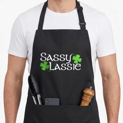 Sassy st patrick day aprons - White Sassy Lassie text with two green shamrocks stacked vertically — shown on a black cotton canvas apron against a white background