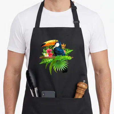 Tropical birds apron - Realistic toucan perched on brown branch with pink flowers and green tropical leaves — shown on a black cotton canvas apron against a white background