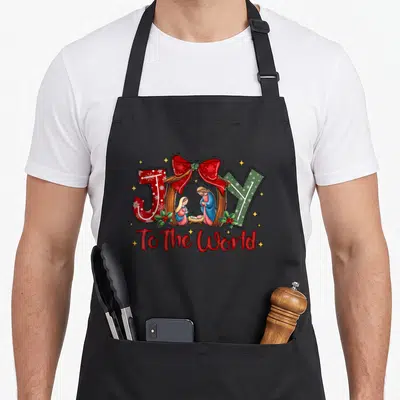 Joy christian womens christmas apron - JOY To The World nativity scene with red bow and stars in cartoon style — shown on a black cotton canvas apron against a white background