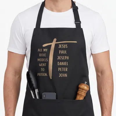Went model apron - Tan cross divides text 'All My Role Models Went to Prison' and biblical names like Jesus Paul — shown on a black cotton canvas apron against a white background