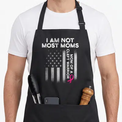 Cleft pink ribbon apron - Distressed USA flag with vertical text I AM NOT MOST MOMS MOM OF A CLEFT WARRIOR and pink — shown on a black cotton canvas apron against a white background