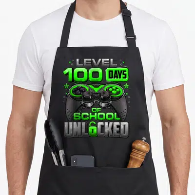 School unlocked day level apron - Game controller with LEVEL 100 DAYS OF SCHOOL UNLOCKED text and two green stars — shown on a black cotton canvas apron against a white background