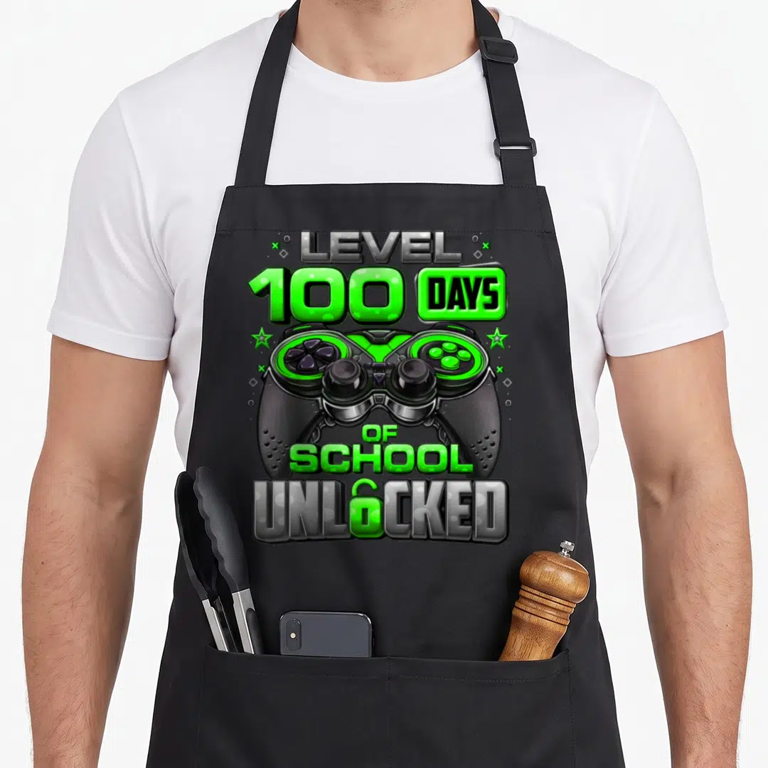 School unlocked day level apron - Game controller with LEVEL 100 DAYS OF SCHOOL UNLOCKED text and two green stars — shown on a black cotton canvas apron against a white background