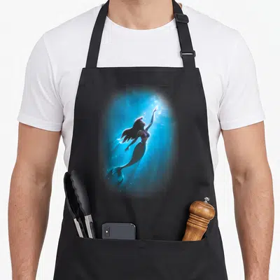 Swim mermaid apron - Realistic mermaid floating diagonally upwards in blue water with light rays and bubbles — shown on a black cotton canvas apron against a white background