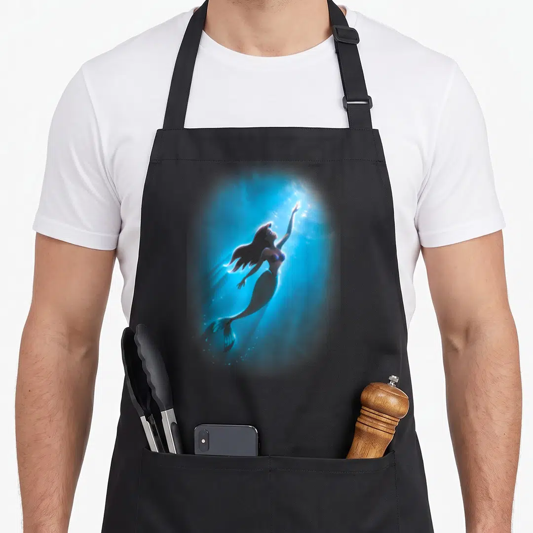Swim mermaid apron - Realistic mermaid floating diagonally upwards in blue water with light rays and bubbles — shown on a black cotton canvas apron against a white background