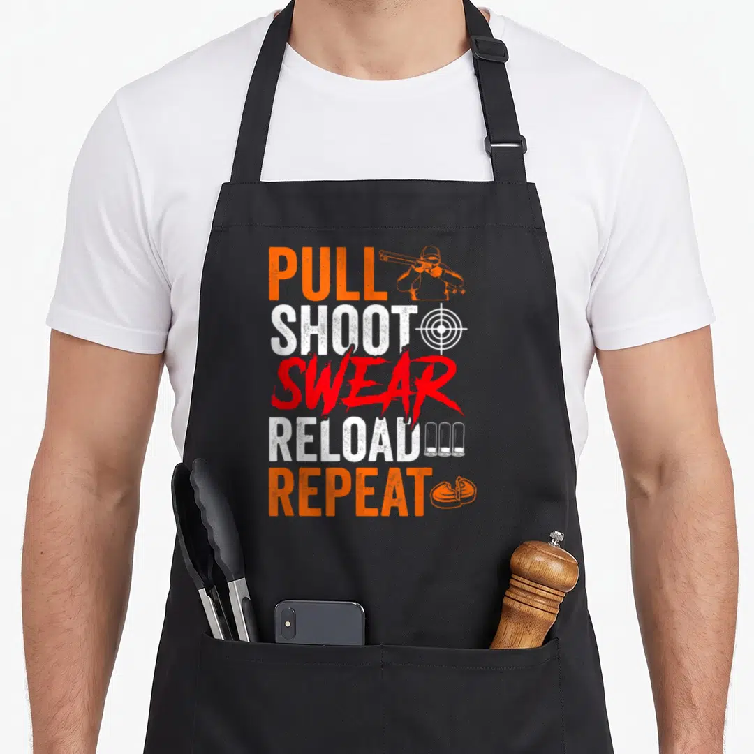 Pull shooting apron - PULL SHOOT SWEAR RELOAD REPEAT text with hunter target casings and earmuff icons in orange white — shown on a black cotton canvas apron against a white background