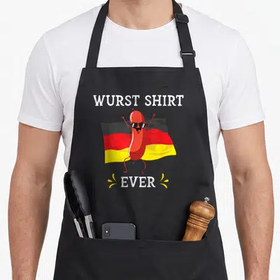 Oktoberfest sausage apron - Cartoon red sausage wearing sunglasses in front of German flag with Wurst Shirt Ever text — shown on a black cotton canvas apron against a white background