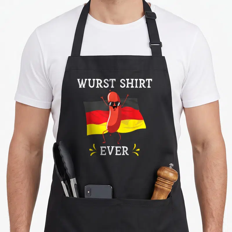 Oktoberfest sausage apron - Cartoon red sausage wearing sunglasses in front of German flag with Wurst Shirt Ever text — shown on a black cotton canvas apron against a white background