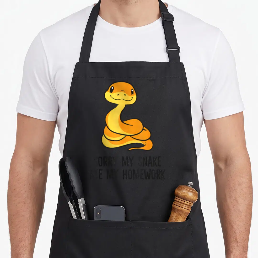 Son pet apron - Cartoon yellow orange snake above sorry my snake ate my homework text — shown on a black cotton canvas apron against a white background