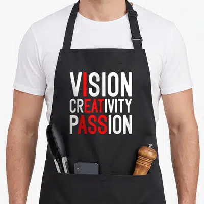 Creativity meme apron - White and red stacked text VISION CREATIVITY PASSION with vertical red column highlighting — shown on a black cotton canvas apron against a white background