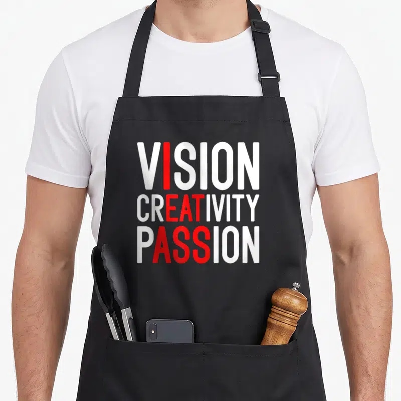 Creativity meme apron - White and red stacked text VISION CREATIVITY PASSION with vertical red column highlighting — shown on a black cotton canvas apron against a white background