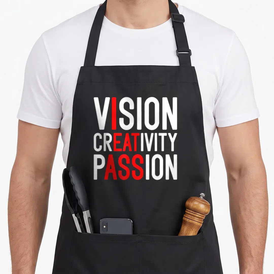 Creativity meme apron - White and red stacked text VISION CREATIVITY PASSION with vertical red column highlighting — shown on a black cotton canvas apron against a white background