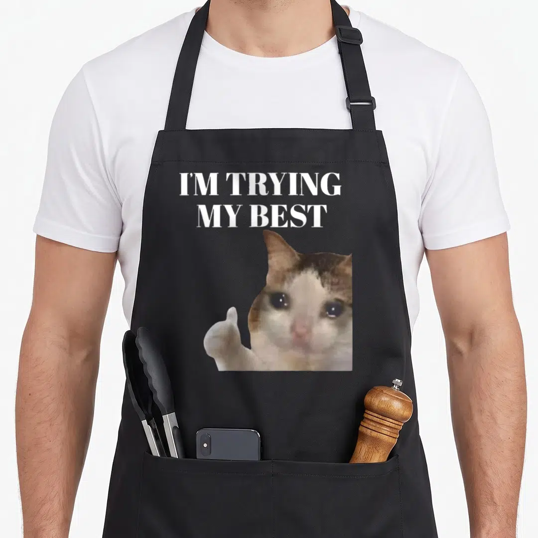 Trying cat apron - Crying cat giving thumbs up with stacked white text I'M TRYING MY BEST on grey background — shown on a black cotton canvas apron against a white background