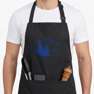 Arawak summer camp chef apron - CAMP ARAWAK text arching over blue oval with trees and sailboat silhouette — shown on a black cotton canvas apron against a white background