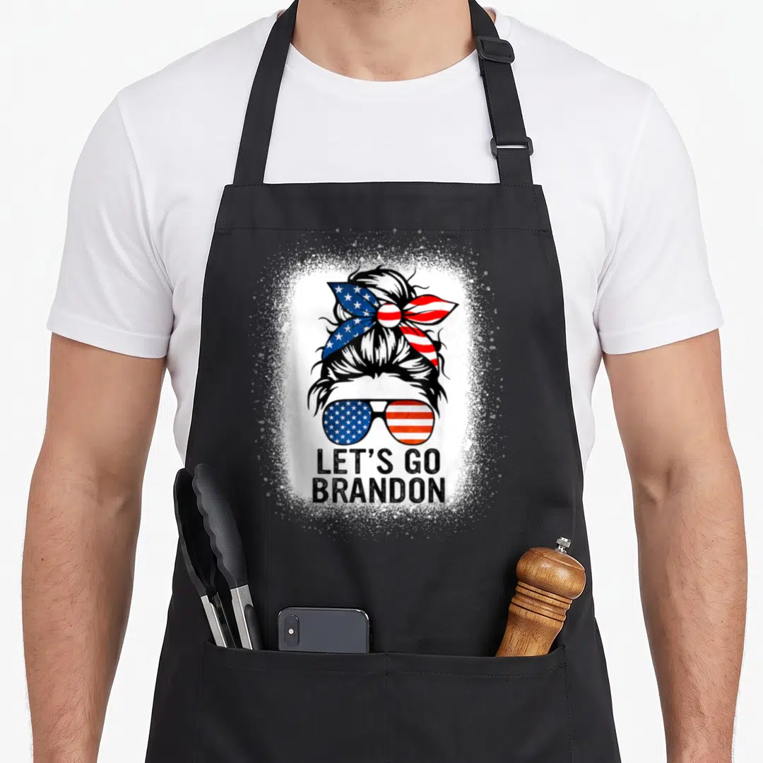 Conservative messy aprons - Patriotic messy bun with flag bandana and sunglasses above Let's Go Brandon text — shown on a black cotton canvas apron against a white background
